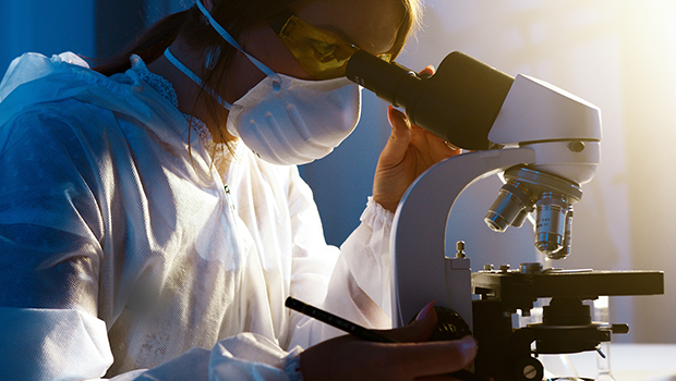 Women in science, an image of a woman staring intently into a microscope, while adjusting the settings on the side of it. She is wearing a white mask, safety glasses and white scrubs but no gloves.